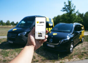 Eine Hand hält ein Smartphone mit BVG-Muva-App. Dahinter stehen zwei Muva-Fahrzeuge.