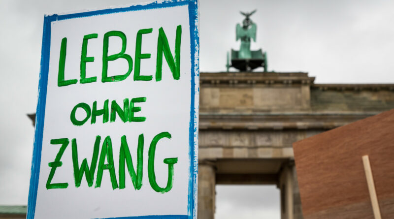 Handgeschriebenes Schild mit grüner Schrift: "Leben ohne Zwang". im Hintergrund das Brandenburger Tor.