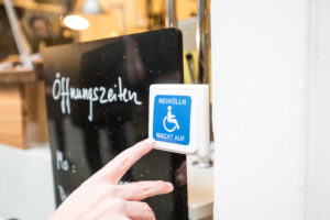 Klingelknopf mit einem blau-weißen Rollstuhl symbol. Links daneben steht hinter einer Glasscheibe eine schwarze Tafel, dort steht Öffnungszeiten drauf.