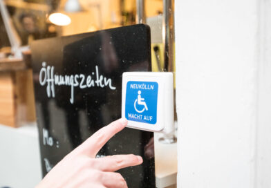 Klingelknopf mit einem blau-weißen Rollstuhl symbol. Links daneben steht hinter einer Glasscheibe eine schwarze Tafel, dort steht Öffnungszeiten drauf.