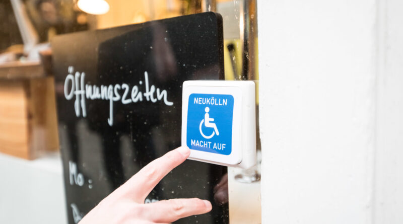 Klingelknopf mit einem blau-weißen Rollstuhl symbol. Links daneben steht hinter einer Glasscheibe eine schwarze Tafel, dort steht Öffnungszeiten drauf.