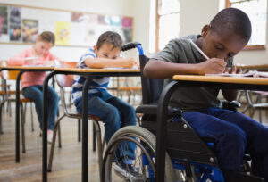 Drei Kinder in der Schule an Tischen. Vorne sitzt ein Kind im Rollstuhl.