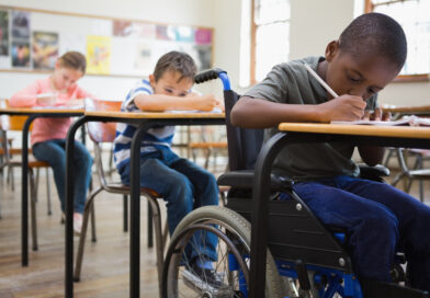 Drei Kinder in der Schule an Tischen. Vorne sitzt ein Kind im Rollstuhl.