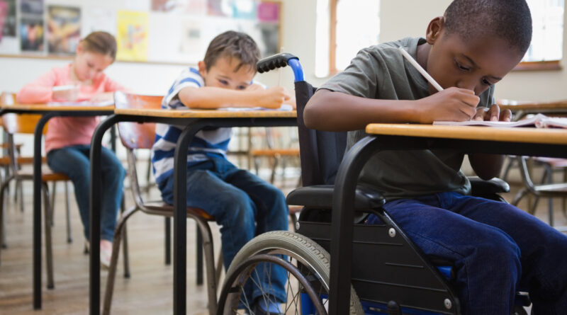 Drei Kinder in der Schule an Tischen. Vorne sitzt ein Kind im Rollstuhl.
